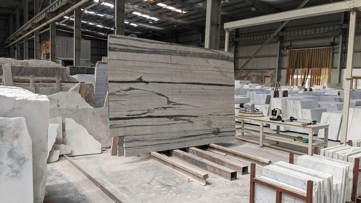 Grey Indian marble slab with dark linear banding in a partner processing hall, beside stacked cut tiles and a polishing machine